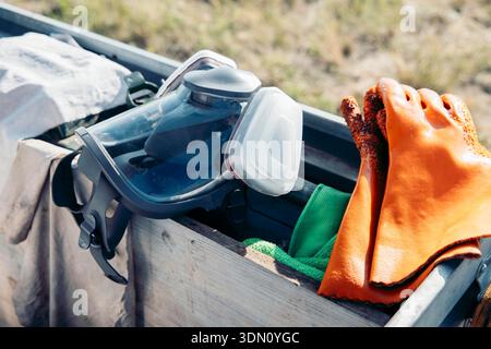 protective equipment stored in wooden box outdoors, safety goggles respirator and orange work gloves arranged, field work in natural environment Stock Photo