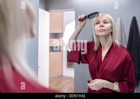 Hair Grooming. Blonde woman with short haircut wearing burgundy silk robe standing in bathroom before mirror while brushing hair Stock Photo