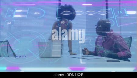 Analyzing, woman in blue top leaning over laptop and man holding papers in boardroom with holograms Stock Photo