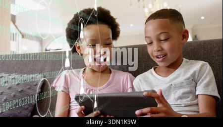 Sitting two children wearing gray tee and pink top, holding tablet in living room with overlays Stock Photo