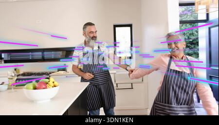 Holding hands, couple wearing matching-striped aprons dancing in kitchen at island with fruit, wine Stock Photo