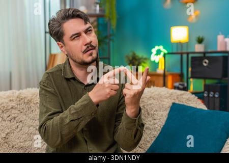 Middle-aged man at home counts tasks on fingers thinking over life choices planning ahead. Young adult guy on sofa ponders decisions hesitates evaluating key goals tracking progress with hand numbers Stock Photo
