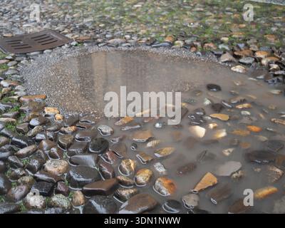 Rainy day puddle reflecting the sky on a cobblestone street with a drain Stock Photo