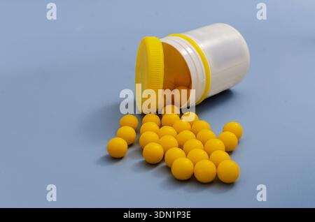 A top-down view of round yellow pills or supplements scattered from an open white bottle with a yellow cap, set against a solid blue background. Stock Photo