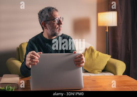 A middle-aged professional man sits at his home office desk, looking away from his laptop to practice mindfulness and take a mental break during a bus Stock Photo