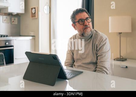 Senior man sitting at a table at home with a tablet, looking away thoughtfully. Concept of mental health, distraction, focus, technology use and quiet Stock Photo