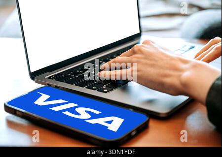 Riga, Latvia - February 04, 2026:Close-up of hands typing on a laptop keyboard with a smartphone on the desk displaying the Visa logo. Concept of onli Stock Photo