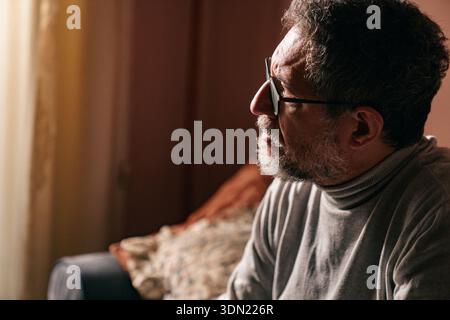 Middle aged man sitting on a sofa at home, resting his face on his hand while thinking. Quiet indoor scene with warm light, expressing contemplation, Stock Photo