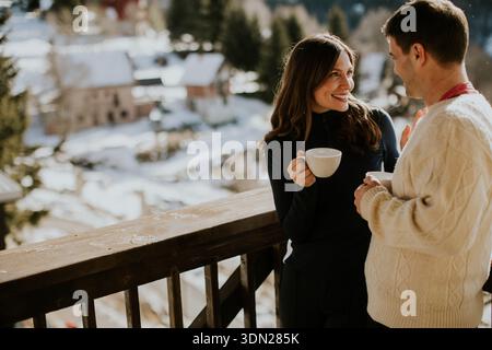 Warm drinks in hand, a couple shares smiles on a cozy mountain balcony as snow blankets the landscape below. Stock Photo