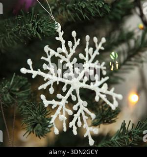 A white snowflake ornament hangs from a green Christmas tree. Background features colorful lights, creating a festive atmosphere Stock Photo