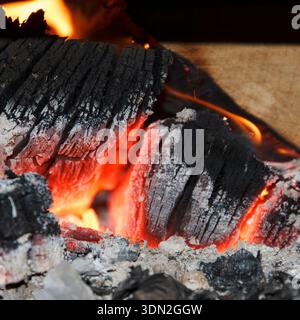 Close-up of glowing embers and ash from a fire. Texture of charred wood is visible, with bright orange and red hues among dark ashes Stock Photo