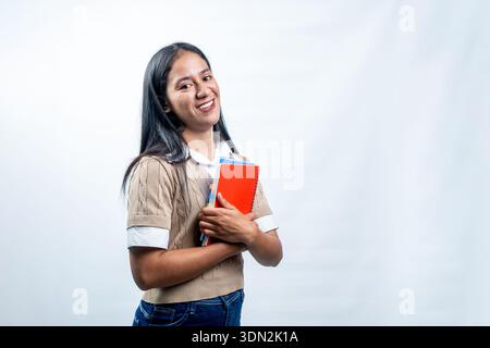 Cheerful young Indonesian female student smiling and holding red notebooks. Concept of education, learning, and university life on white background. Stock Photo