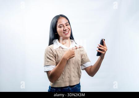 Cheerful young Indonesian woman smiling and pointing at her phone. Concept of mobile app recommendation, digital marketing, or online shopping. Stock Photo