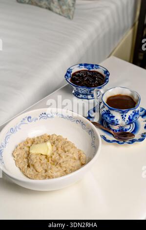 Breakfast in bed with oatmeal, a serving of jam and hot coffee. A bowl of oatmeal with butter next to a coffee pair and jam on a bedside table in a ho Stock Photo