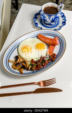 Fried eggs with sausages, mushrooms and beans on a plate next to coffee in a hotel room. A hearty breakfast with fried eggs, sausages and black coffee Stock Photo