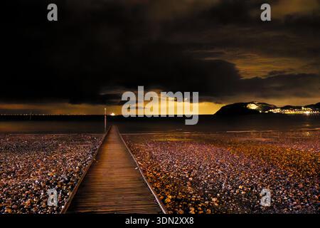 A wooden walkway crosses a pebbly beach to a dark sea with an eerie glow in the post sunset sky and black silhouetted headland, Llandudno, North Wales. Stock Photo