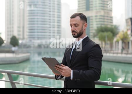 Confident businessman stands by a waterway in Dubai using a digital device while surrounded by skyscrapers. Stock Photo