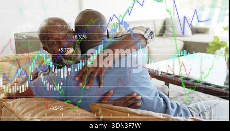 Embracing father-son wearing plaid denim shirts on tan sofa in living room, with financial charts Stock Photo