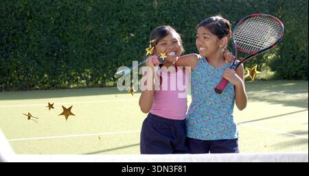 Smiling two girls in tank tops standing by net on tennis court, holding rackets, gold stars Stock Photo