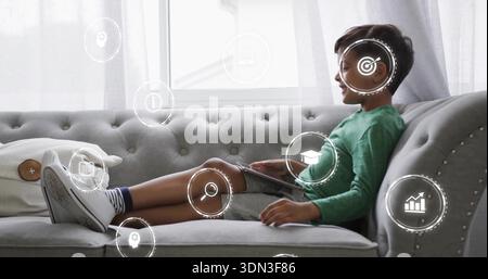 Reclining boy in green shirt, sneakers on tufted sofa at home, holding tablet with digital icons Stock Photo