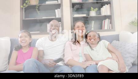 Smiling family sitting on sofa at home, man holding TV remote, bookcases behind, girls in dresses Stock Photo