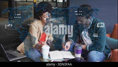Leaning pair in blazer and cardigan reviewing notes in lounge, with laptop, coffee and data overlay Stock Photo