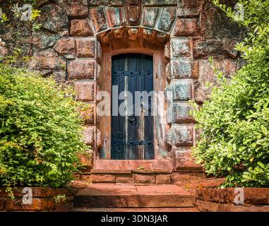 Ancient dark wooden door with ornate iron hinges set in a rustic stone archway surrounded by lush green bushes Stock Photo