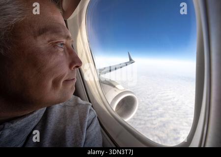 Adult male airline passenger gazes through an aircraft window at wing and clouds under a deep blue sky, enjoying the view during flight. Travel and aviation lifestyle concept Stock Photo
