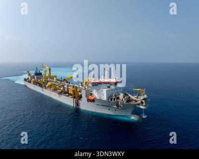 Dhidhdhoo, Maldives - 30 January 2026: Aerial view of a large grey dredger ship cutting through the deep blue sea, leaving a trail of light blue sedim Stock Photo