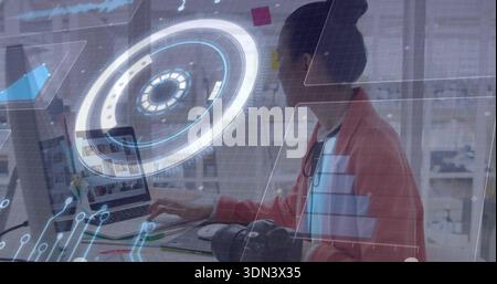 Working mid-adult Asian woman typing on laptop at modern studio, red jacket, holographic UI visible Stock Photo