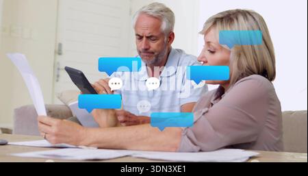 Reviewing couple in blue polo and blouse checking tablet and papers at home, chat bubbles Stock Photo
