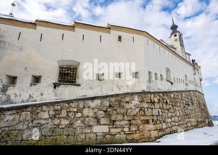 A white plaster fortress wall sits atop a rugged stone base, with small windows and a distant church tower. Snow covers the ground beneath a clear blu Stock Photo