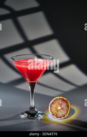 Martini glass filled with a vibrant red drink placed next to a blood orange slice, casting a colorful light halo Stock Photo