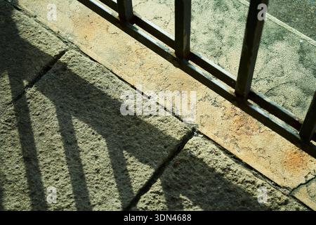 High contrast sunlight casting geometric shadows from a metal railing onto a textured old stone pavement, creating a minimalist abstract pattern. Stock Photo