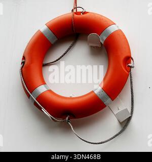 Bright orange lifebuoy ring with reflective bands and attached rope, mounted on a white nave wall. The ropes are securely fastened through four eyelet Stock Photo