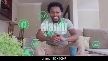Sitting man in green tee and khakis leaning forward in living room, smiling with eco icons Stock Photo