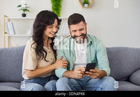 Happy couple using smartphone together at home, sitting on sofa in living room Stock Photo