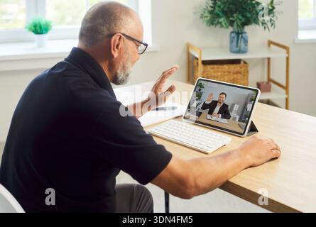 Senior man waving during online videocall on tablet at bright home office Stock Photo