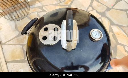 A top-down view of a black barbecue grill lid featuring a sturdy handle and an adjustable air vent, set on an outdoor terrace. Stock Photo
