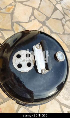 A top-down view of a black barbecue grill lid featuring a sturdy handle and an adjustable air vent, set on an outdoor terrace. Stock Photo