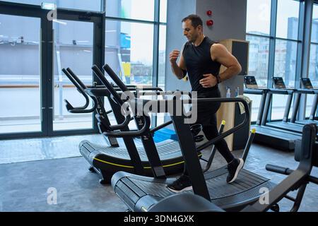 Middle-aged man running on curved treadmill for cardio workout at modern gym with large windows Stock Photo