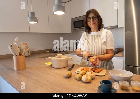 Smiling mature woman wearing apron peeling carrot while preparing vegetables in modern kitchen. Healthy homemade food, everyday lifestyle and cozy dom Stock Photo