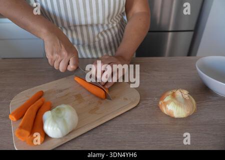 Carrot being cut with a knife on a wooden cutting board while preparing vegetables in a home kitchen. Selective focus and natural light. Stock Photo