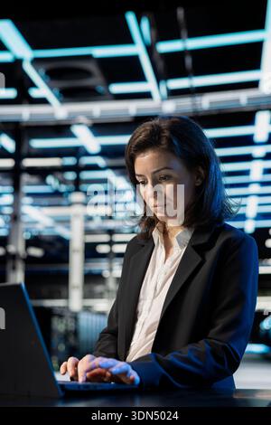 Data center female technician at desk writing code on laptop, updating equipment network systems. Woman in server room typing on notebook keyboard, coding to upgrade supercomputers Stock Photo