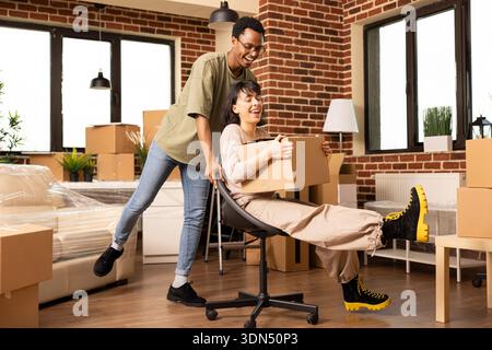 African american boyfriend pushing caucasian girlfriend on chair through unfurnished living room, enjoying thrill of fresh start together. Cheerful couple laughing and bonding during moving day. Stock Photo