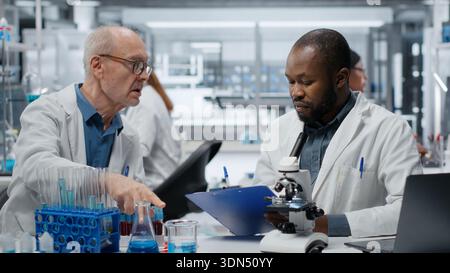 Medical team in lab conducting research on blood sample, taking notes. African american man and elderly colleague in laboratory analyzing sanguine fluid specimen, writing down findings, camera B Stock Photo