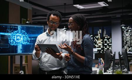 Tech agency staff members using tablet, brainstorming how to optimize AI performance in office. African american technicians use machine learning on device for debugging, analyzing code Stock Photo