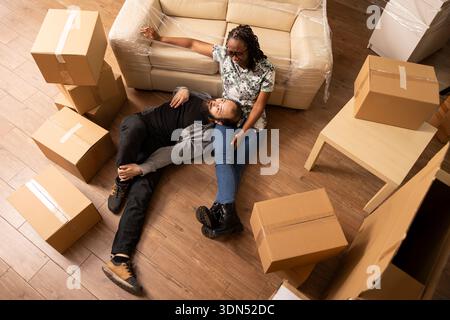 Caucasian boyfriend lying on lap of black girlfriend in living room filled with packed cardboard boxes. Diverse couple sits on floor, taking break during relocation day while waiting for moving truck. Stock Photo