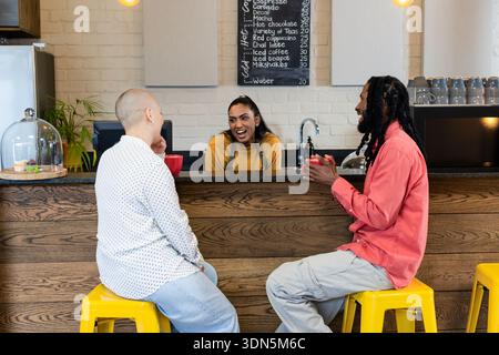 Indian barista and two customers smiling and interacting at cafe counter while holding red mugs Stock Photo