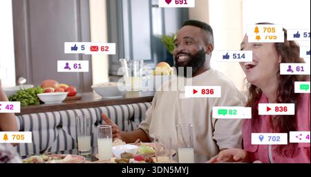 Laughing man in cream top and woman in pink chatting at dining table, with plates, notifications Stock Photo